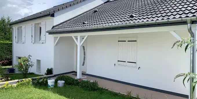 Maison blanche récemment rénovée avec toit tuilé foncé, volets blancs. Porche couvert, jardin verdoyant et deux pots de peinture au sol.