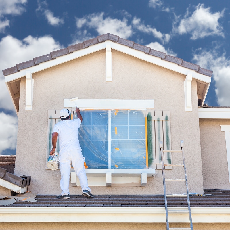 Un peintre en blanc utilise un rouleau pour repeindre la façade d'une maison beige avec une fenêtre protégée. Échelle à côté, ciel nuageux.