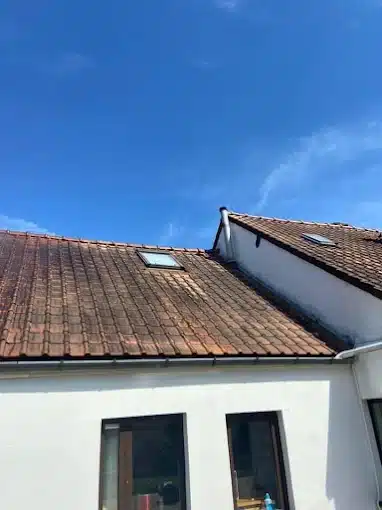 Toit de maison en tuiles rouges tachées de saleté, avec deux lucarnes, sous un ciel bleu. Mur blanc et fenêtres visibles.
