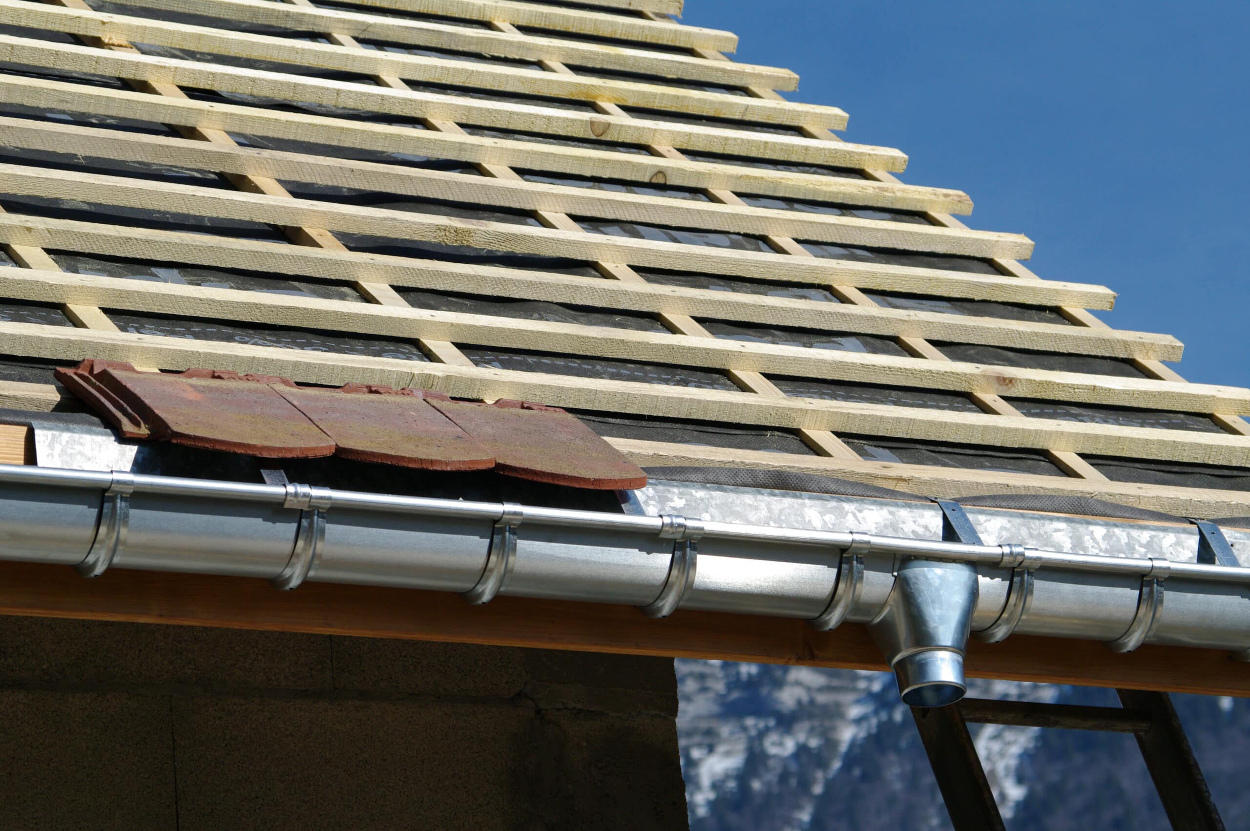 Structure de toit en construction: lattes en bois sur membrane noire, gouttière métallique neuve, quelques tuiles en terre cuite, ciel bleu.