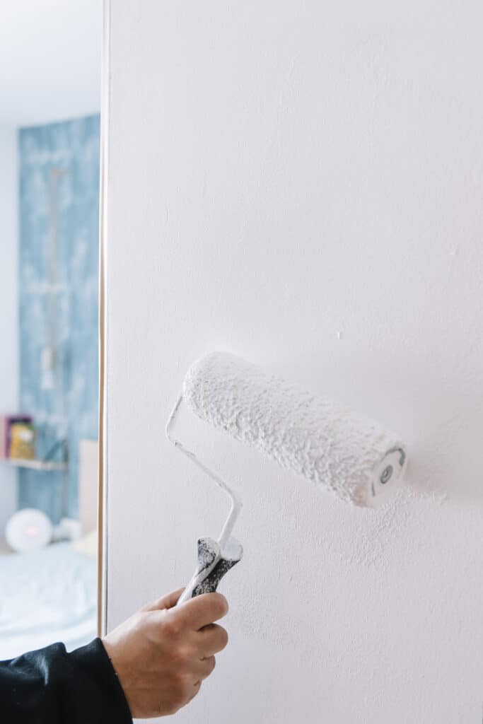 Une main tient un rouleau de peinture, appliquant de la peinture blanche sur un mur texturé. Arrière-plan flou d'une pièce.