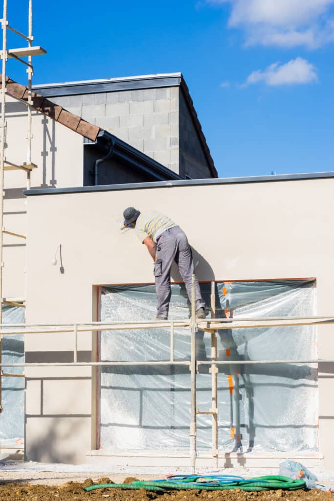 Ouvrier sur échafaudage appliquant l'enduit beige sur la façade d'une maison en construction sous un ciel bleu clair.